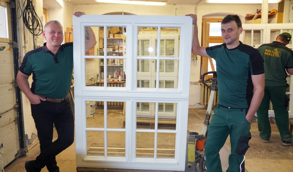 Auf dem Bild sind zwei Mitarbeitende des Unternehmens Holz Hess zu sehen. Sie halten jeweils rechts und links ein Fenster mit weißem Rahmen.